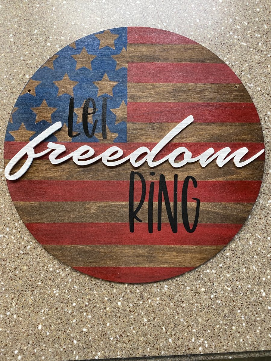 Round wood door sign painted with an American flag design in red, white, and blue with layered wood 'Let Freedom Ring' lettering