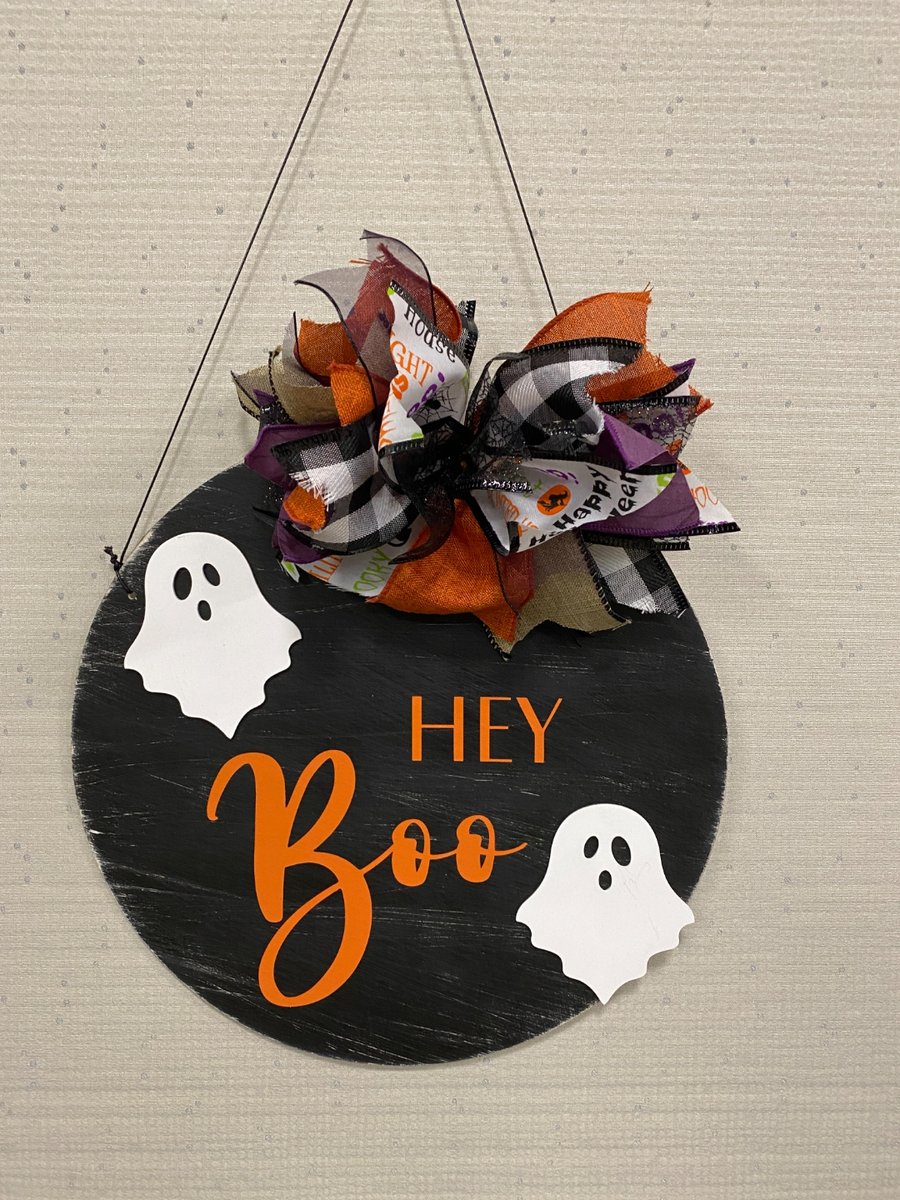 Round black wood door hanger painted with 'Hey Boo' in orange lettering and two white ghost designs, topped with a large Halloween ribbon bow